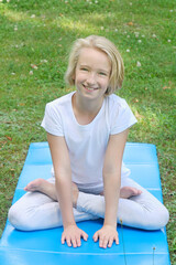 Beautiful and happy blonde kid girl of school age practices yoga on the mat in the park. Outdoor warm-up, healthy lifestyle. 