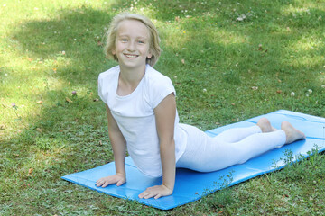 Beautiful and happy blonde kid girl of school age go in for sports and doing stretching on the mat in the park. Outdoor warm-up, healthy lifestyle. 