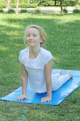 Beautiful and happy blonde kid girl of school age go in for sports and doing stretching on the mat in the park. Outdoor warm-up, healthy lifestyle. 