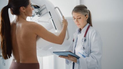 Friendly Female Doctor with Tablet Computer Explains Mammogram Procedure to a Topless Adult Female...