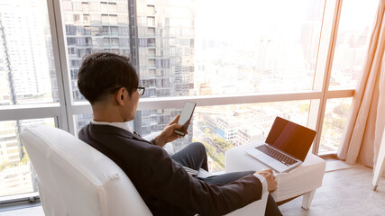 Asian businessman in smart suit and holding smart phone and looking at it while sitting at his working place in the hotel after seminar