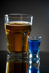 Premade setup for saint patrick drink. Shot glass of blue curacao drink next to the light gold beer. Reflecting surface and dark gradient background