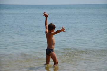 Fototapeta na wymiar A little boy with his hands up stands in the sea. A happy child stands on a sea beach. An enthusiastic child on the seashore. The concept of happiness, freedom, joy, rest, pleasure.