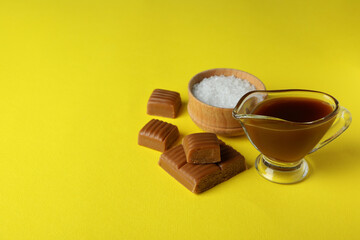 Caramel pieces, sauce and bowl of salt on yellow background