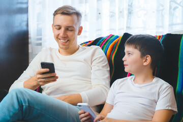 Cheerful young family dad son toddler childwatching online video on smartphone sitting on couch together, laughing parent kids texting playing games entertaining using mobile phone apps home