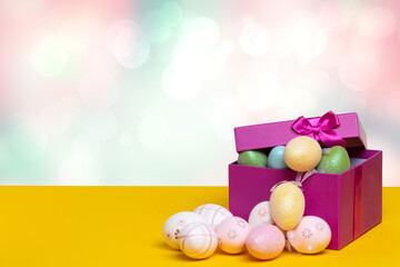 Happy Easter greeting card template. Close up of colourful Easter eggs and an open gift box with eggs on yellow table against abstract blurred pastel background. Copy space for design.