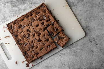 Dark chocolate brownie cut into 16 square pieces. The item is on white plastic cutting board with chocolate crumbs