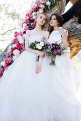two brides with bouquets are sitting on the background of a decorated tree in a meadow