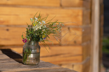 Modest bouquet of white and pink wildflowers with green twigs in glass jar is placed on wooden table near wall of house outside on blurred natural background. Summer still life. Pleasant memories.
