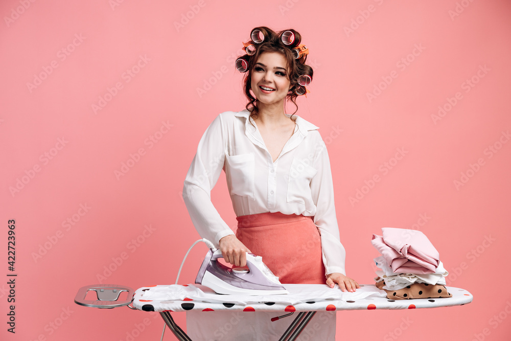 Wall mural Attractive young female housewife ironing clean clothes while doing household chores, isolated on pink background. Cleaning concept.