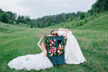 two girls in wedding dresses with bouquets are sitting on a meadow
