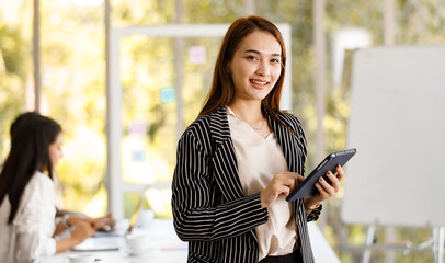 portrait of young beautiful Asian businesswoman with long hair wearing smart casual holding tablet while smiling to a camera with soft background of meeting room and a colleague