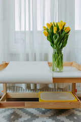 Bright fresh yellow tulips on white background. Bunch of yellow tulips in big glass jar in white interior. Spring flowers in glass vase on white table at home.