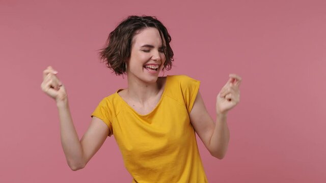 Short hairdo young woman 20s in yellow t-shirt dance clenching fists waving rising expressive gesticulate hands fool around have fun celebrate play fluttering hair isolated on pastel pink background
