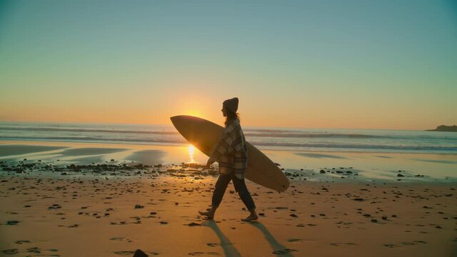 Side shot of young woman in cool casual outfit walk with surfboard at empty epic sandy beach. Millennial and cinematic authentic real surfer female. Surfer lifestyle and copy space 
