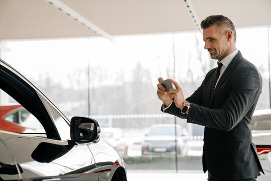 Mature man taking photo on cellphone while choosing car in showroom