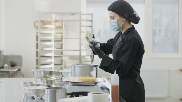 Side view of young woman in Covid-19 face mask putting dessert topping in confectionery syringe as Caucasian man entering commercial kitchen. Professional confectioners cooking pastry indoors.