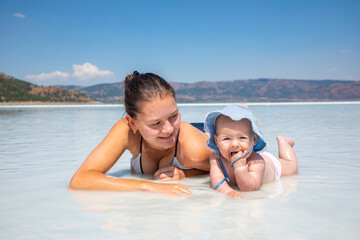 Young woman in swim suit and her daughter little baby girl are laying on white clay of turquoise crater lake Salda Golu, Turkey