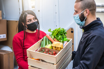 Young woman courier delivers to customer man a basket of fruit and vegetables bought online at supermarket wearing face mask and gloves during the global Coronavirus Covid-19 pandemic