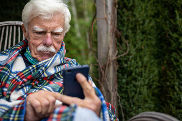 Senior man sitting in the rocking chair reading text message on smartphone. Mature man using his smartphone on the veranda. Portrait of a retired man reading a message on his cellular.