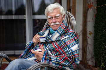 An older man wrapped in a blanket relaxes on a porch in the garden, drinking coffee.