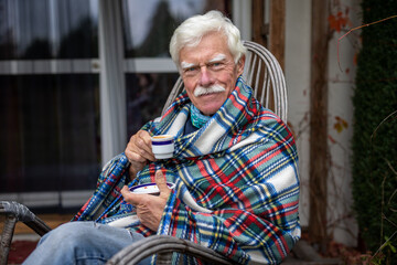 An older man wrapped in a blanket relaxes on a porch in the garden, drinking coffee.