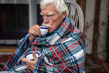 An older man wrapped in a blanket relaxes on a porch in the garden, drinking coffee.