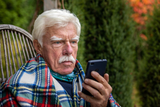 An Elderly Gentleman Is Having A Phone Call. Senior Man Sitting In The Rocking Chair During His Smartphone Conversation. Mature Man Using His Cellular On The Veranda.