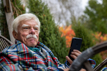 An elderly gentleman is having a phone call. Senior man sitting in the rocking chair during his smartphone conversation. Mature man using his cellular on the veranda.