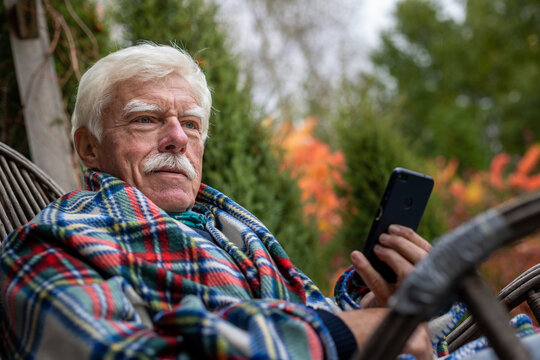 An Elderly Gentleman Is Having A Phone Call. Senior Man Sitting In The Rocking Chair During His Smartphone Conversation. Mature Man Using His Cellular On The Veranda.