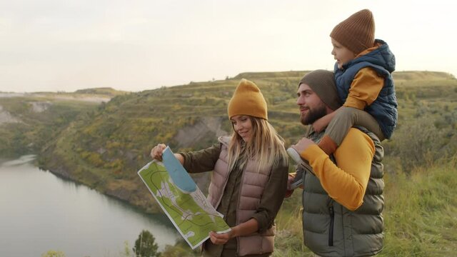 Handheld slowmo of family of three standing on green hill overlooking lake. Cute 5-year-boy sitting on shoulders of his bearded father and pointing at scenery while cheerful mother looking at map