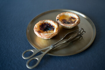 Two delicious Belem cakes (Pasteis de Belem) on a bronze tray, with serving tongs.  Portuguese...