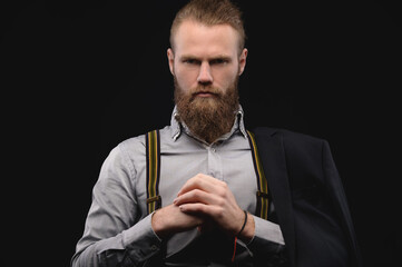 Portrait of an attractive Caucasian bearded man with a stylish hairstyle in a shirt with suspenders sitting in a dark room. Shows a gesture of strength
