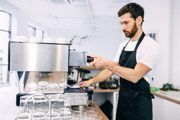 A professional, experienced barista prepares coffee in a coffee machine, wearing a black apron and white T-shirt.