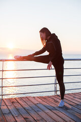 caucasian woman is stretching near a handrail on the embankment at sunrise near a sea