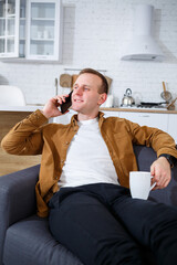 Happy young man is sitting on the sofa in the living room with a cup of coffee and talking on the phone.