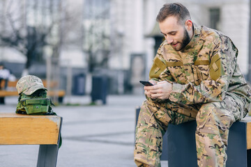 A military soldier sits on a bench at a bus stop waiting for a bus and looks at the phone and smiles