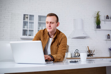 An attractive young man in casual clothes sitting in the kitchen using a laptop computer. Work from home, remote workflow.