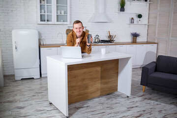 Handsome young businessman working with computer remotely sitting at the table in the kitchen. A pleasant happy person communicates on a social network, searches for information on the Internet.