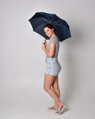 full length portrait of girl wearing shirt and denim shorts, holding an umbrella. isolated on grey studio background.