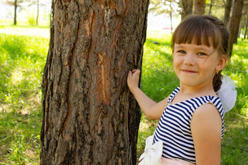 A little beautiful girl in a dress stands near the trunk of a tree in the pine forest