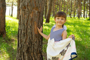 A little beautiful girl in a dress stands near the trunk of a tree in the pine forest