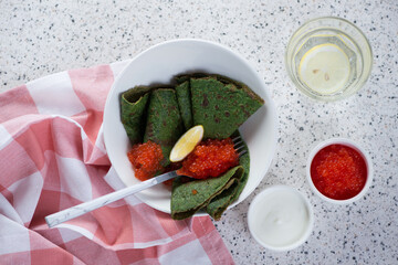 Spinach crapes with red caviar and sour cream over beige stone background, top view, horizontal shot