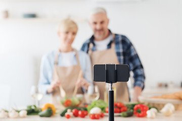 Smiling middle aged man and lady preparing salad and looking at smartphone webcam in kitchen