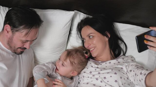 Happy young family with a baby girl, are photographed on a smartphone in the bed. Mother, father and daughter-bloggers in the room communicate using a modern gadget online. Modern young family