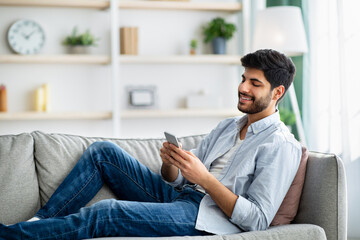 Mobile communication concept. Happy arab man using cellphone, browsing internet, sitting on couch...