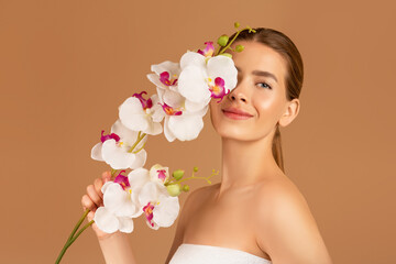 Beautiful millennial woman touching her face with orchid on brown studio background
