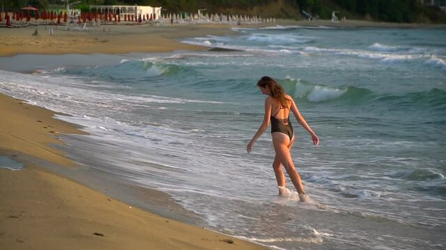 Beautiful happy woman on the beach in a swimsuit. A young girl runs along the ocean under the rays of the sun. Merry vacation at Kazat and dawn with sea waves. High quality FullHD footage
