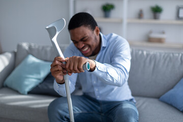 Young black man suffering from acute pain, leaning on crutch at home, selective focus