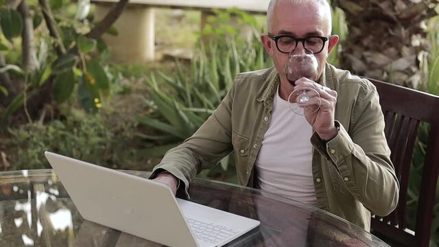 Middle age man sitting outdoors by the table and working on laptop. Man drink dry red wine during the work on backyard. Freelancer on quarantine